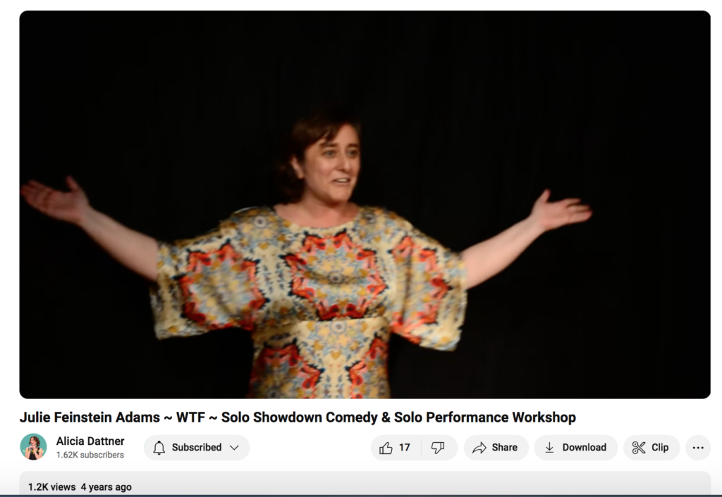 Performer onstage, light skin, dark hair, in mandala-print silk shirt with black background, arms outstretched and sort of smiling while telling a story about her spouse's gender transition